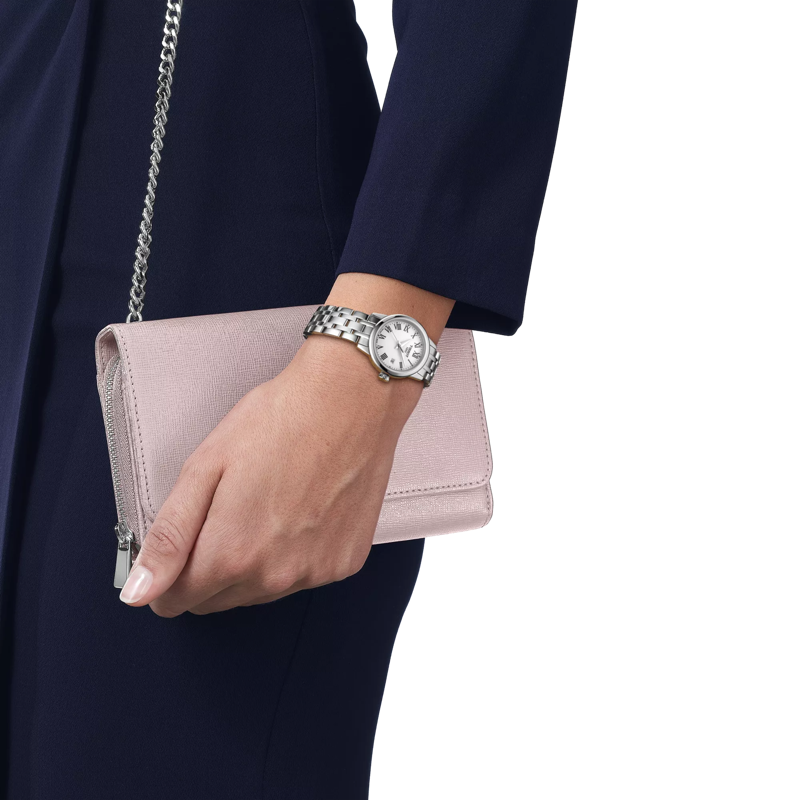 Woman wearing Tissot Ladies Dream watch with roman numeral dial and steel bracelet, holding pink clutch.