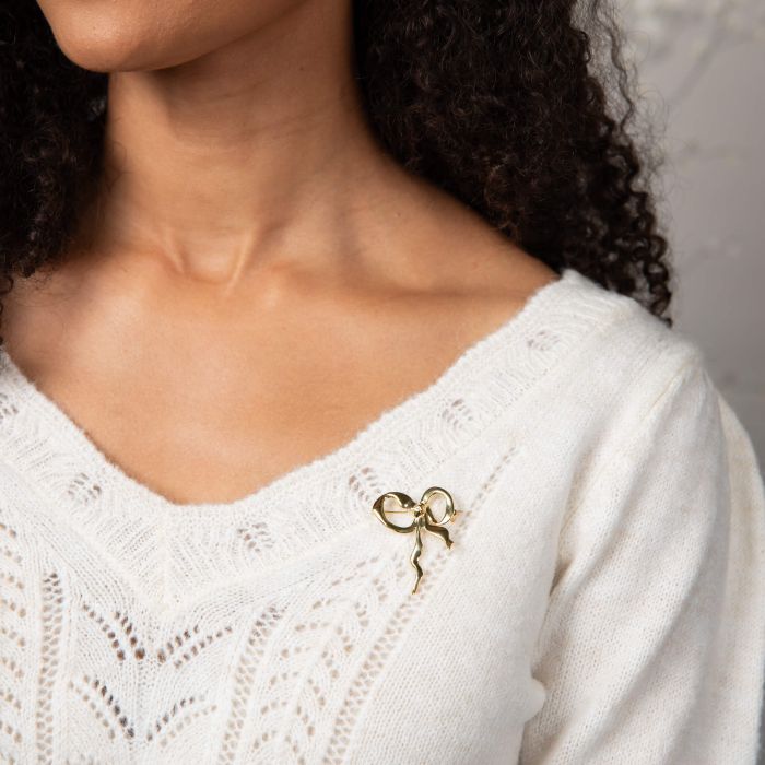 Woman wearing Laura Ashley statement ribbon bow brooch, gold plated, displayed on white knit sweater.