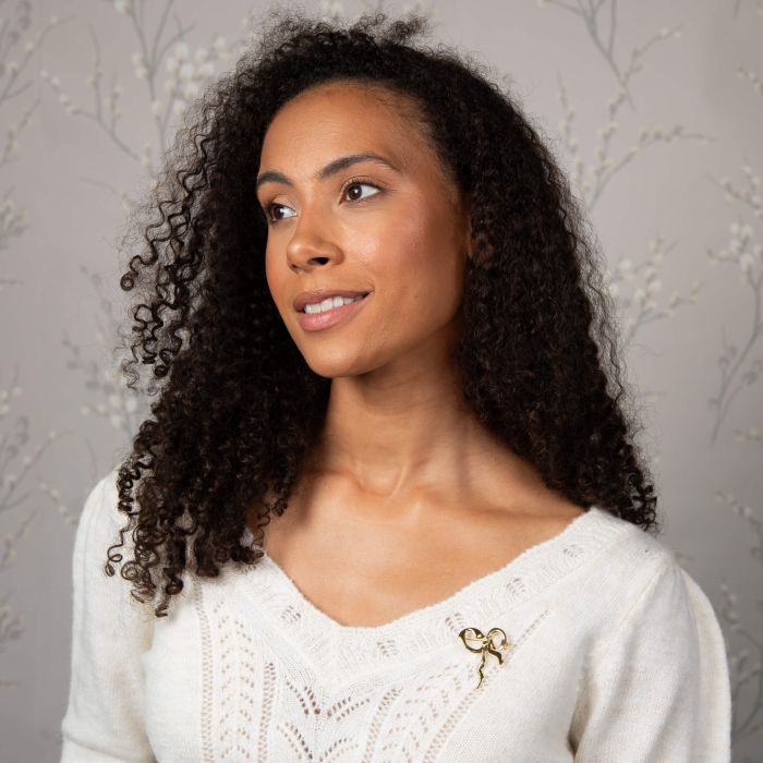 Woman wearing Laura Ashley gold-plated ribbon bow brooch on a white v-neck sweater. Elegant jewelry accessory.